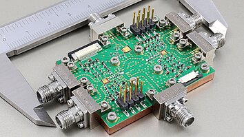 Photograph shows the demonstrator module with the full-duplex topology, comprising two power amplifiers and a low-noise amplifier combined with a printed circuit board, which is mounted onto a copper heat sink. The module is placed between the jaws of a caliper for size reference.