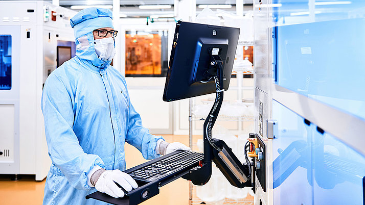 A laboratory technician in protective clothing is working on a computer. He is wearing a protective mask and gloves. The screen is on a stand, while laboratory equipment is visible in the background. 