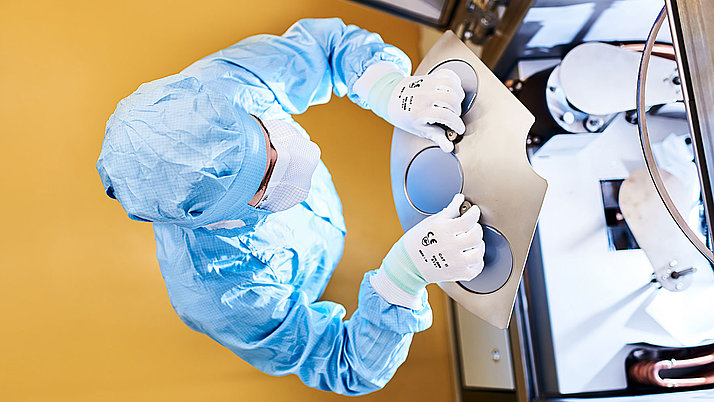 Eine Person in einem blauen Schutzanzug und Handschuhen arbeitet an einem technischen Gerät. Sie hält eine Metallplatte mit zwei Öffnungen, während sie präzise Handgriffe ausführt.