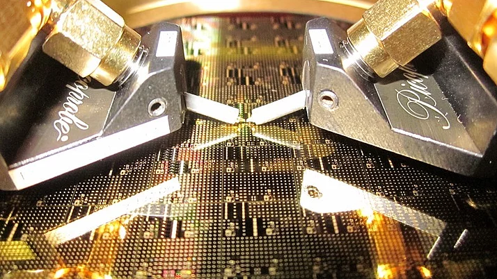 Close-up view of two metallic probes positioned above a patterned circuit board. The probes are connected to gold-plated connectors. The circuit board features a complex design.