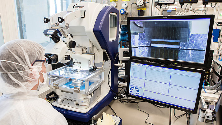 Ein Wissenschaftler trägt Schutzkleidung und arbeitet an einem Mikroskop. Auf dem Bildschirm sind vergrößerte Details einer Probe zu sehen.