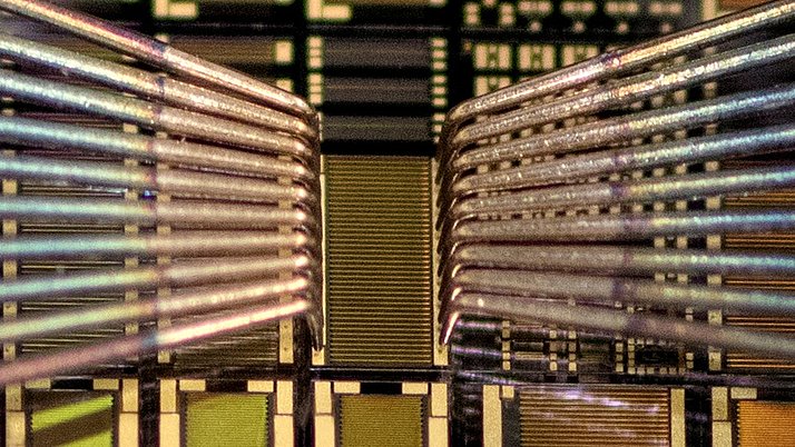 Nahaufnahme von elektrischen Leitungen und Halbleiterstrukturen auf einem Mikrochip. Die feinen Drähte verlaufen parallel und zeigen eine komplexe Anordnung von goldenen und grünen Schaltkreisen.