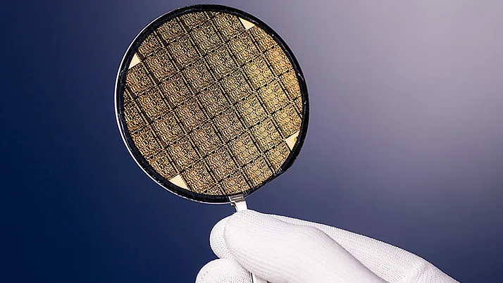 A hand in a white glove holds a circular silicon wafer, showcasing a grid-like pattern. The wafer's surface has a textured, metallic sheen.