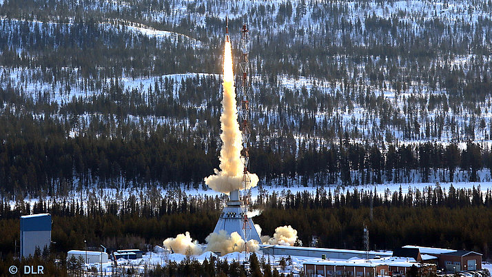 Eine Rakete startet von einer Startplattform, wobei ein kräftiger Schub von Rauch und Dampf entsteht. Im Hintergrund sind schneebedeckte Wälder und Hügel zu sehen.