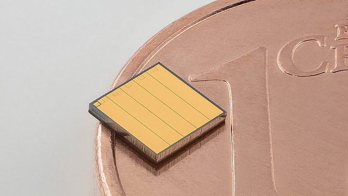 A small, square microchip with a gold surface is positioned on a copper penny. The chip has rectangular sections and is slightly elevated above the penny's surface.