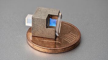 A small, square object with a rough, metallic surface, with a blue lens protruding from the right and a white lens protruding from the left. It is an optical isolator placed on a one-cent coin.