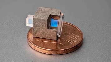 Miniaturisierter optischer Isolator: ein kleiner, rau strukturierter Würfel mit zwei gegenüberliegenden transparenten rechteckigen Prismen, von denen eines einen bläulich schimmernden Innenbereich zeigt, platziert auf einer Ein-Cent-Münze.