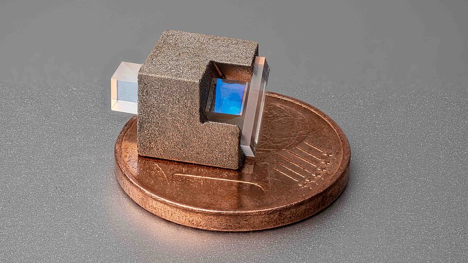 Miniaturisierter optischer Isolator: ein kleiner, rau strukturierter Würfel mit zwei gegenüberliegenden transparenten rechteckigen Prismen, von denen eines einen bläulich schimmernden Innenbereich zeigt, platziert auf einer Ein-Cent-Münze.