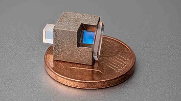 Miniaturisierter optischer Isolator: ein kleiner, rau strukturierter Würfel mit zwei gegenüberliegenden transparenten rechteckigen Prismen, von denen eines einen bläulich schimmernden Innenbereich zeigt, platziert auf einer Ein-Cent-Münze.