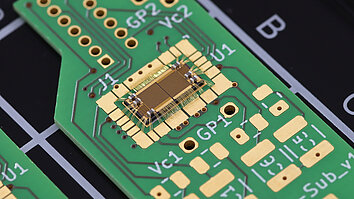 Das Bild zeigt eine Leiterplatte mit grüner Grundfarbe. In der Mitte ist ein kleiner, goldfarbener Chip, eine Halbbrücke mit inegrierten Gate-Treibern  sichtbar, umgeben von goldenen und silbernen Lötpunkten. Es gibt auch mehrere beschriftete Kontaktstellen auf der Platine.