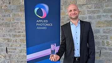 A man in a suit is holding an award and smiling. In the background is a banner that reads “APPLIED PHOTONICS AWARD.”