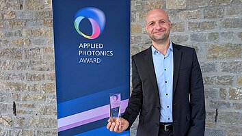 A man in a suit is holding an award and smiling. In the background is a banner that reads “APPLIED PHOTONICS AWARD.”