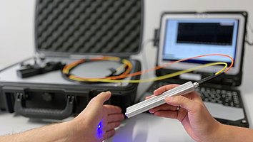 Raman-Sensor-System auf einem Tisch zusammen mit einem Monitor und dem Koffer. Bestandteil ist auch eine Optode, aus deren Spitze bläuliches Licht kommt.