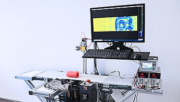 Terahertz-Linienscanner im Betrieb. Auf dem Laufband liegt eine Kunststoffrolle, in die ein Tesafilmroller eingewickelt ist. Dieser lässt sich auf dem darüber liegenden Monitor erkennen.