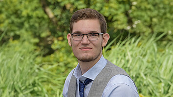 Ein junger Mann mit Brille trägt ein graues Weste über einem hellblauen Hemd und einer blauen Krawatte. Er lächelt und sitzt vor einer grünen, bewachsenen Kulisse.