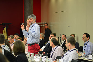 A group of people are sitting at tables in a conference room. A man with gray hair and a blue shirt stands and speaks while the other participants listen attentively. 