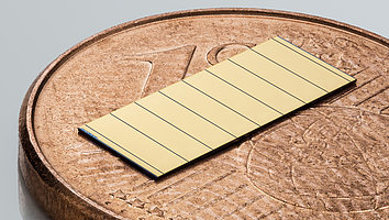 Das Bild zeigt einen goldfarbenen Laserbarren mit einer glatten Oberfläche, der auf einer 1-Cent-Münze liegt. Der Laserbarren ist ein dünnes, rechteckiges Bauteil mit mehreren dunklen Linien, die seine Segmente abgrenzen. Die 1-Cent-Münze dient als Größenvergleich und verdeutlicht die geringe Größe des Laserbarrens. 