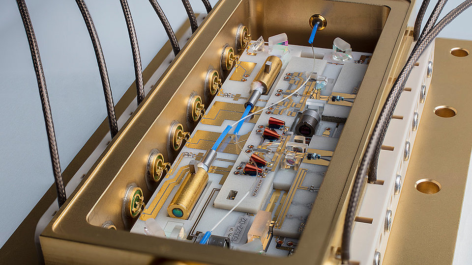 An intricate electronic circuit housed in a gold-colored casing. The circuit includes various components such as cylindrical connectors, coils, and optical elements.