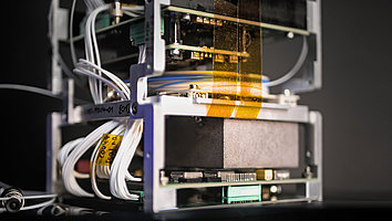 Close-up of a payload of a nanosatellite with quantum experiments. Electrical components and cables are visible, as well as a gray box. This contains the FBH's highly compact laser system. The background is dark, which draws attention to the details of the equipment.
