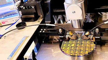 Semiconductor wafer with patterned circuits mounted on a test stage, contacted by probe needles beneath a microscope and surrounded by laboratory measurement equipment.