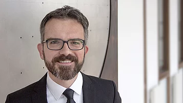Ein Mann mit Brille und Bart trägt einen Anzug und lächelt. Der Hintergrund zeigt unscharfe Wände in einem modernen Gebäude. Der Mann strahlt Freundlichkeit und Professionalität aus.