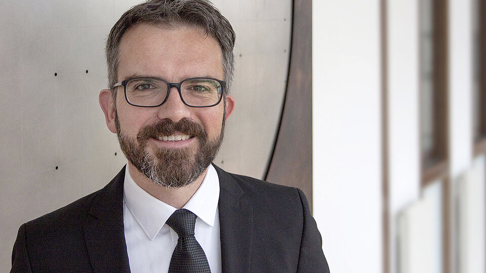 Ein Mann mit Brille und Bart trägt einen Anzug und lächelt. Der Hintergrund zeigt unscharfe Wände in einem modernen Gebäude. Der Mann strahlt Freundlichkeit und Professionalität aus.