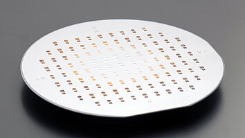 Round semiconductor wafer viewed at an angle, patterned with evenly spaced small square and circular cells in rows across its surface.