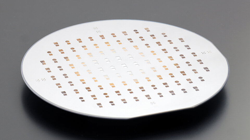 Round semiconductor wafer viewed at an angle, patterned with evenly spaced small square and circular cells in rows across its surface.