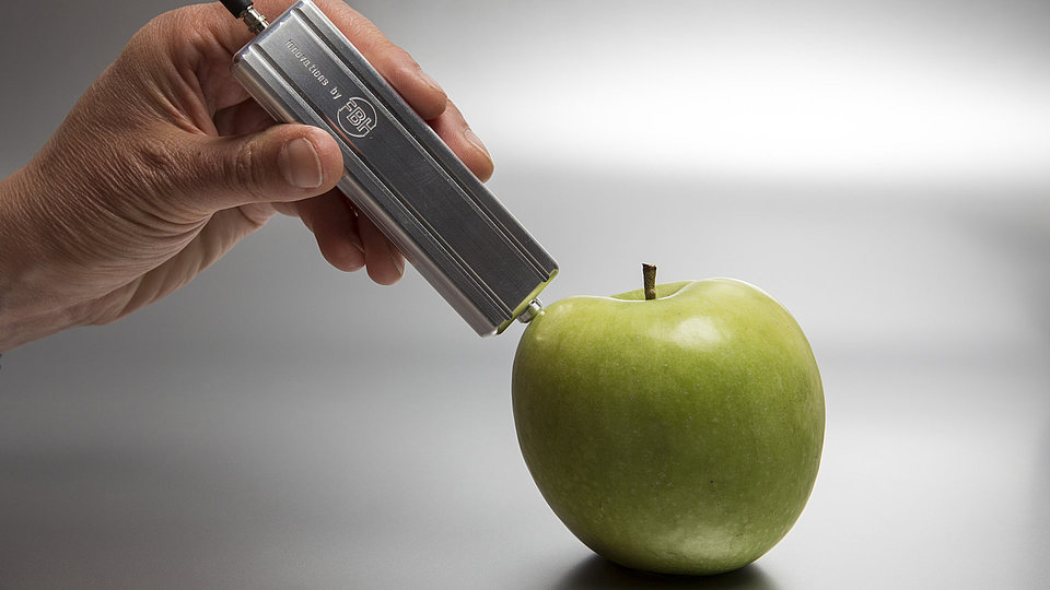 A hand holds a miniaturized measuring head to an apple. This optode is suitable for spectroscopic measurements.