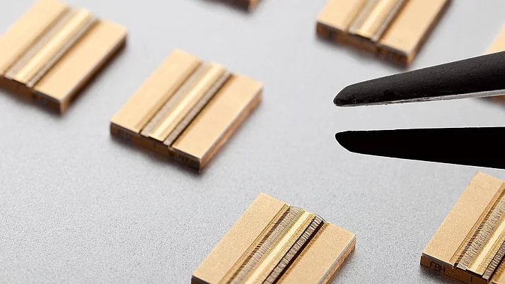 Nahaufnahme von goldfarbenen elektronischen Bauteilen, die auf einer grauen Oberfläche liegen. Eine Pinzette hält ein Bauteil, während andere Bauteile im Hintergrund sichtbar sind. 