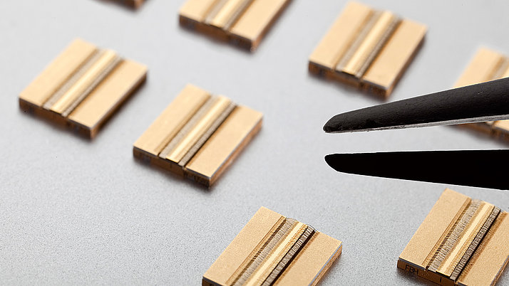 Close-up of gold-colored electronic components lying on a grey surface. Tweezers hold a component while other components are visible in the background. 