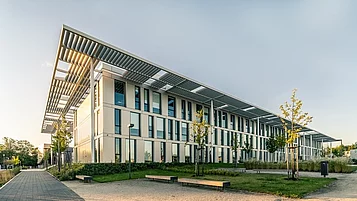 Ein modernes, zweigeschossiges Gebäude mit großen Fenstern und einem überstehenden Dach. Der Eingang wird von jungen Bäumen und einer grünen Fläche umgeben.
