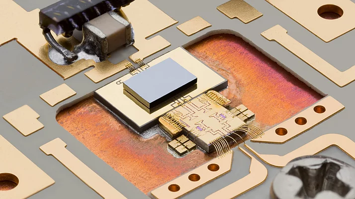 Close-up of a microchip on a circuit board, showcasing intricate wiring and components. The chip is centrally located with a shiny metallic surface, surrounded by golden paths and connectors on a patterned substrate.