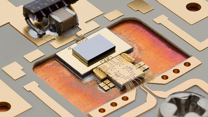Close-up of a microchip on a circuit board, showcasing intricate wiring and components. The chip is centrally located with a shiny metallic surface, surrounded by golden paths and connectors on a patterned substrate.