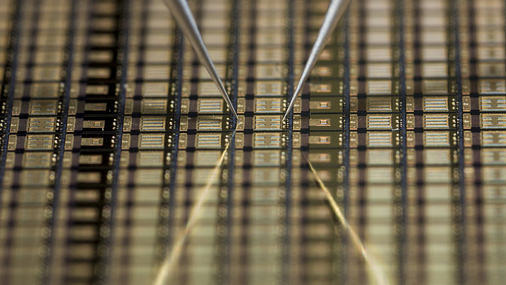 Silicon wafer with finely structured electrical circuits. Two fine tweezers hold the wafer in the center while it rests on a reflective surface.