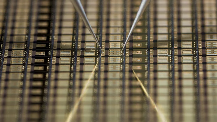 Silicon wafer with finely structured electrical circuits. Two fine tweezers hold the wafer in the center while it rests on a reflective surface.