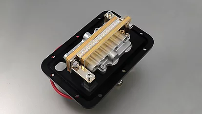 Ein mechanisches Bauteil auf einem schwarzen Grund, mit einem silbernen Gehäuse und einer goldenen Leiste. Das Teil enthält verschiedene bewegliche Elemente und Anschlüsse.