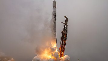 Eine Rakete startet aus einer Startrampe, während Flammen und Rauch aus ihren Triebwerken austreten. Der Himmel ist bewölkt und die Szene vermittelt ein Gefühl von Bewegung und Energie.