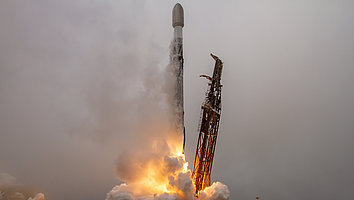 A rocket launches from a launch pad as flames and smoke pour from its engines. The sky is cloudy, and the scene conveys a sense of movement and energy.