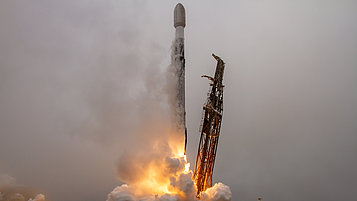 A rocket launches from a launch pad as flames and smoke pour from its engines. The sky is cloudy, and the scene conveys a sense of movement and energy.