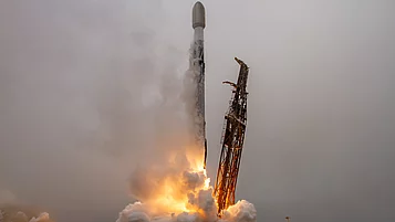 Eine Rakete startet aus einer Startrampe, während Flammen und Rauch aus ihren Triebwerken austreten. Der Himmel ist bewölkt und die Szene vermittelt ein Gefühl von Bewegung und Energie.