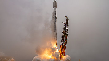 Eine Rakete startet aus einer Startrampe, während Flammen und Rauch aus ihren Triebwerken austreten. Der Himmel ist bewölkt und die Szene vermittelt ein Gefühl von Bewegung und Energie.