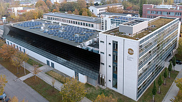 An aerial view of a modern building with solar panels on the roof, featuring a combination of glass and metal facades. The surrounding area has greenery and parking spaces.
