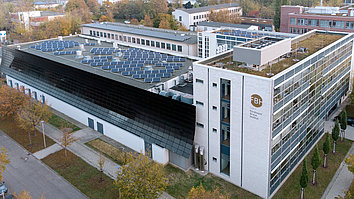 Modernes Gebäude mit einer Mischung aus Glas- und Metallfassade. Auf dem Dach befinden sich Solarpanels. Um das Gebäude wachsen Bäume und es gibt gepflegte Grünflächen sowie Parkplätze daneben.