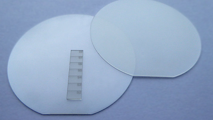 Two transparent, round wafers lie next to each other on a surface. Next to them is a thin glass scale with test lines numbered from S2 to S7. 
