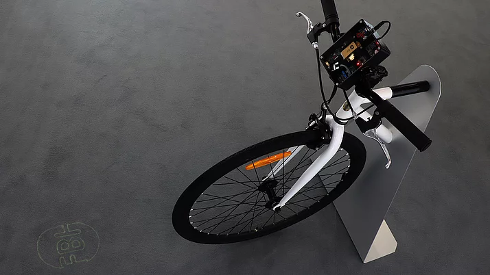 Ein modernes Fahrrad mit einem Steuerungselement am Lenker. Das Fahrrad hat einen weißen Rahmen und wird von einem stabilen, grauen Ständer gehalten.
