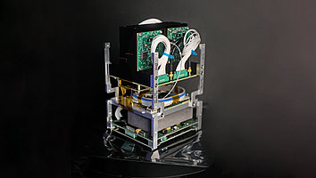 Payload of a nanosatellite carrying quantum experiments. It consists of several layers of circuit boards, cables, and a black cover on top. A gray box is also visible. It contains the FBH's highly compact laser system. The components are structured and connected to a frame. The background is dark, which draws attention to the instrument.