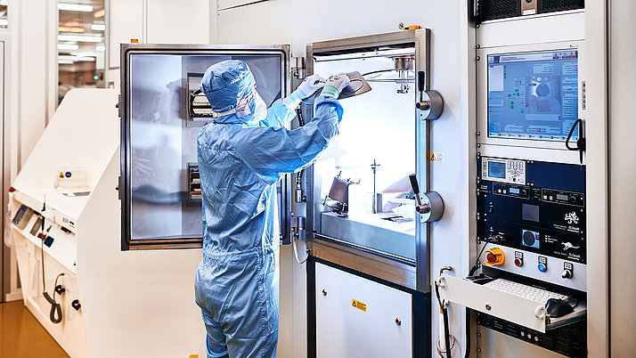 A person in a cleanroom suit is carefully handling equipment inside a high-tech apparatus. The environment shows various controls and displays visible on a nearby console.