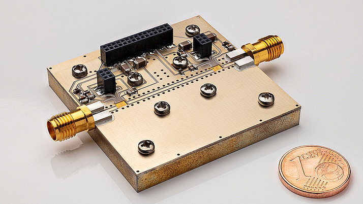Close-up of a rectangular electronic circuit board featuring metallic connectors. Two gold-colored connectors are prominently positioned on opposite sides of the board. A one euro cent coin is placed for scale.