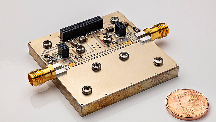 Close-up of a rectangular electronic circuit board featuring metallic connectors. Two gold-colored connectors are prominently positioned on opposite sides of the board. A one euro cent coin is placed for scale.